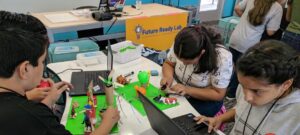 Children working with computers are models. The models have wires coming out which are attached to microcontrollers. A sign in the back reads "Future Ready Lab"