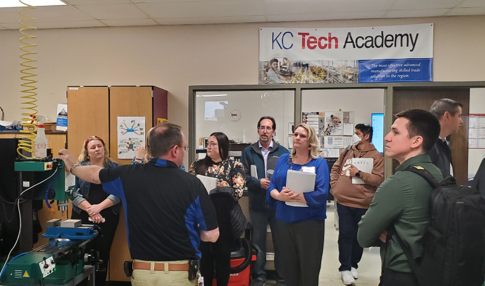 A group of teachers is gathered in a classroom. One person, leading the group, has his hand on a large piece of machinery. In the background a sign hangs reading "KC Tech Academy"