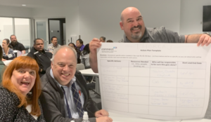 Three smiling people are featured in the foreground, one holds a large printout with the ConnectED logo. It says "Thoreaua [Highschool]" at the top, and is filled in with handwritten information. They are in a conference room filled with other people.